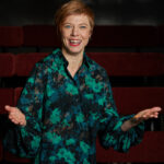 Porträtt av kursledaren Karin Bergstrand som gestikulerar med båda händerna. Hon ler och är iförd en lång grön mönstrad skjorta.